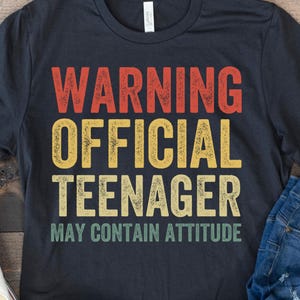 Puede incluir: Una camiseta azul marino con el texto "WARNING OFFICIAL TEENAGER MAY CONTAIN ATTITUDE" en rojo, amarillo y verde. La camiseta se combina con vaqueros azules y zapatillas blancas sobre una superficie de madera.