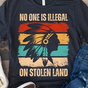 May include: A black t-shirt featuring a vintage-style graphic of a Native American wearing a headdress. The graphic is set against a background of red, orange, yellow, green, and blue stripes. The text "No one is illegal on stolen land" is printed in white on the shirt.