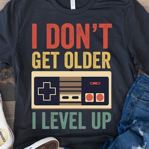 Könnte beinhalten: Dunkelblaues T-Shirt mit dem Text "I DON'T GET OLDER I LEVEL UP" und einer Retro-Videospiel-Controller-Grafik. Das Shirt wird mit blauen Jeans und weißen Turnschuhen kombiniert.