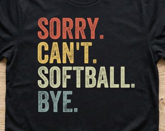 May include: A black short-sleeved t-shirt featuring the text SORRY. CAN'T. SOFTBALL. BYE. printed in a stacked, distressed font. The words are coloured in a gradient of red, yellow, light green, and light blue against the dark fabric.