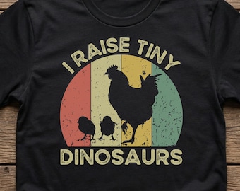 May include: Black t-shirt with a retro design featuring a hen and chicks silhouette inside a colorful circle. The text reads "I RAISE TINY DINOSAURS" in a vintage font, perfect for a farm or animal lover.