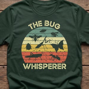 May include: Dark green t-shirt featuring a retro design with various insect silhouettes inside a circular shape. The text "THE BUG WHISPERER" is arched above and below the insect illustrations, which include a butterfly, ant, and grasshopper.
