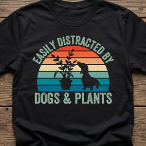 Op de afbeelding: Een zwart T-shirt met korte mouwen en een afbeelding van een kamerplant en het silhouet van een hond tegen een retro zonsondergang met oranje, gele en blauwe strepen. De tekst luidt EASILY DISTRACTED BY DOGS & PLANTS in een lichtgroen lettertype.