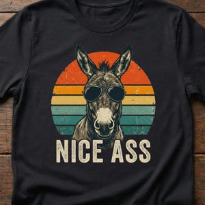 May include: Black t-shirt featuring a donkey wearing sunglasses, set against a retro sunset design. The text "NICE ASS" is printed in white below the donkey. The shirt is made of a soft material.