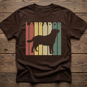 May include: Brown t-shirt featuring a retro design with the word "LABRADOR" in a vertical striped pattern of red, yellow, green, and cream colors. A silhouette of a Labrador dog is centered within the stripes.