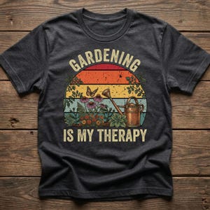 May include: Dark gray t-shirt featuring a colorful graphic with the text "Gardening is My Therapy." The design includes a sunset, flowers, a butterfly, and a watering can, perfect for gardening enthusiasts.