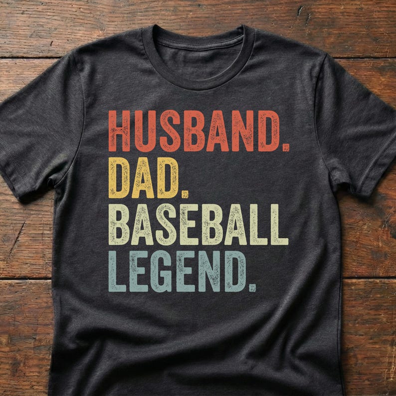 A flat lay mockup of a dark heather grey t-shirt featuring bold, colorful, and distressed athletic block lettering that reads 'HUSBAND. DAD. BASEBALL LEGEND.' A perfect baseball dad shirt, casual game day apparel, and funny Father's Day gift for a husband or team coach.