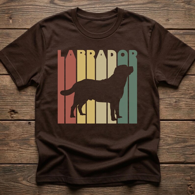 May include: Brown t-shirt featuring a retro design with the word "LABRADOR" in a vertical striped pattern of red, yellow, green, and cream colors. A silhouette of a Labrador dog is centered within the stripes.