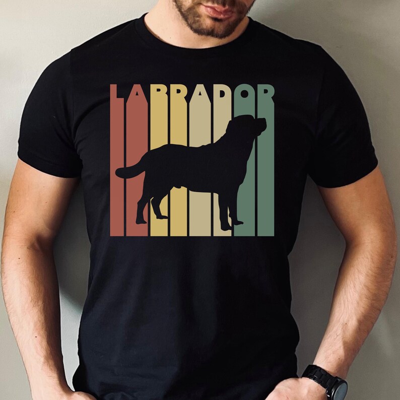 May include: Black t-shirt featuring a Labrador silhouette with the word "LABRADOR" above. The design includes vertical stripes in shades of red, yellow, and green, creating a retro aesthetic. The shirt is made of a soft material.