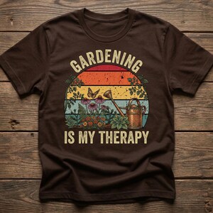 May include: Brown t-shirt with a colorful graphic. The graphic features a sunset design with the words "Gardening is my Therapy" in white. The design includes flowers, a watering can, and a butterfly.