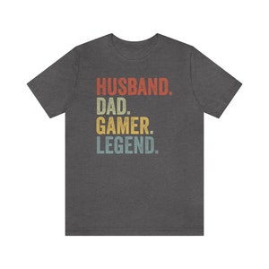 May include: Dark gray t-shirt with a vintage-style graphic that reads "Husband. Dad. Gamer. Legend."