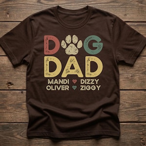 May include: A brown t-shirt with the words "DOG DAD" in colorful, distressed lettering. A paw print is between the words. Below the words are names with heart symbols. A casual, comfortable tee.
