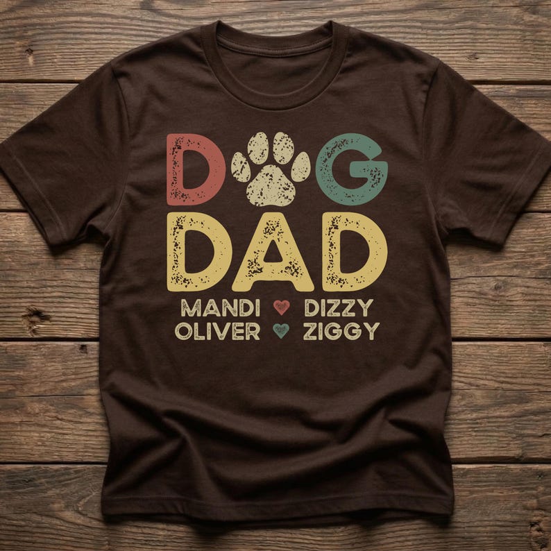 May include: A brown t-shirt with the words "DOG DAD" in colorful, distressed lettering. A paw print is between the words. Below the words are names with heart symbols. A casual, comfortable tee.