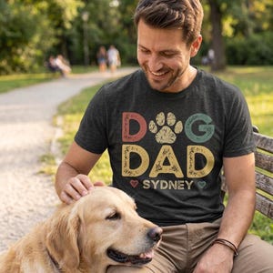 May include: Dark gray t-shirt with the text "Dog Dad Sydney" in a colorful, distressed font. The design includes a paw print and hearts. A man is smiling while petting a golden retriever.