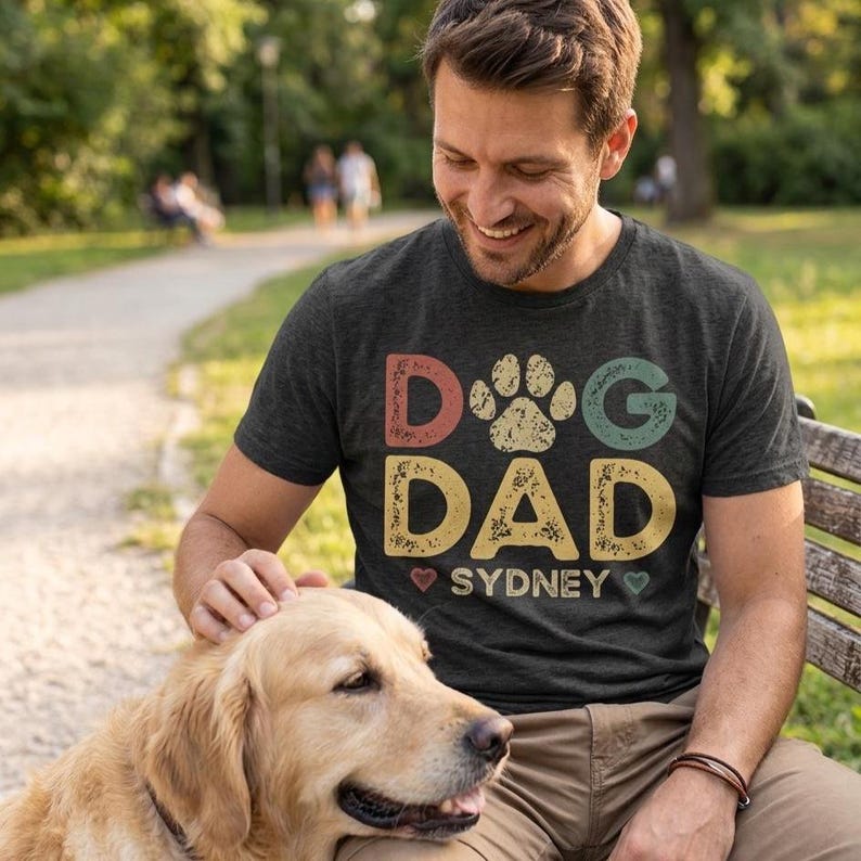 May include: Dark gray t-shirt with the text "Dog Dad Sydney" in a colorful, distressed font. The design includes a paw print and hearts. A man is smiling while petting a golden retriever.