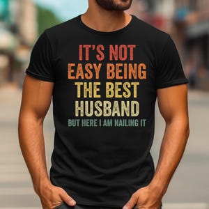 May include: Black t-shirt with the text "IT'S NOT EASY BEING THE BEST HUSBAND BUT HERE I AM NAILING IT" in orange, yellow, and white lettering. The shirt is a crew neck and has short sleeves.