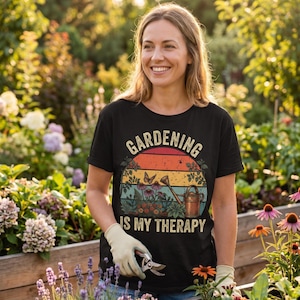 May include: Black t-shirt with the text "GARDENING IS MY THERAPY" in a vintage design. The graphic includes a sunset, butterflies, flowers, and a watering can. The person is wearing gardening gloves and holding pruning shears.