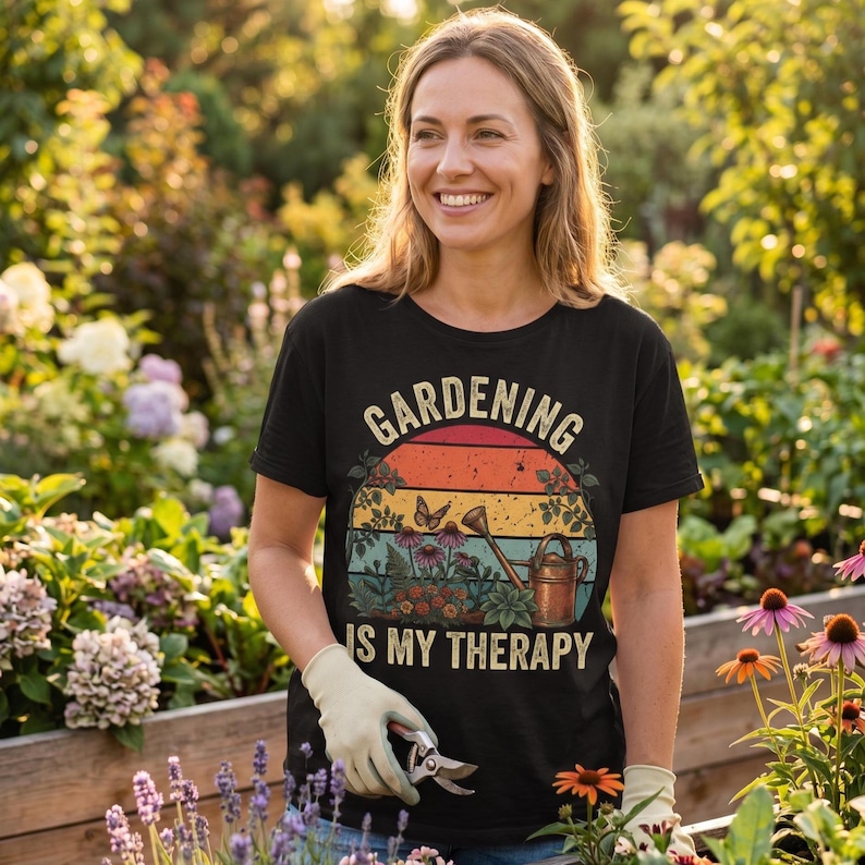 May include: Black t-shirt with the text "GARDENING IS MY THERAPY" in a vintage design. The graphic includes a sunset, butterflies, flowers, and a watering can. The person is wearing gardening gloves and holding pruning shears.