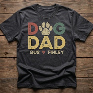 May include: Dark gray t-shirt with the words "DOG DAD" in colorful, distressed lettering, a paw print, and the names "Gus" and "Finley" with a heart symbol. The shirt is on a wooden surface.