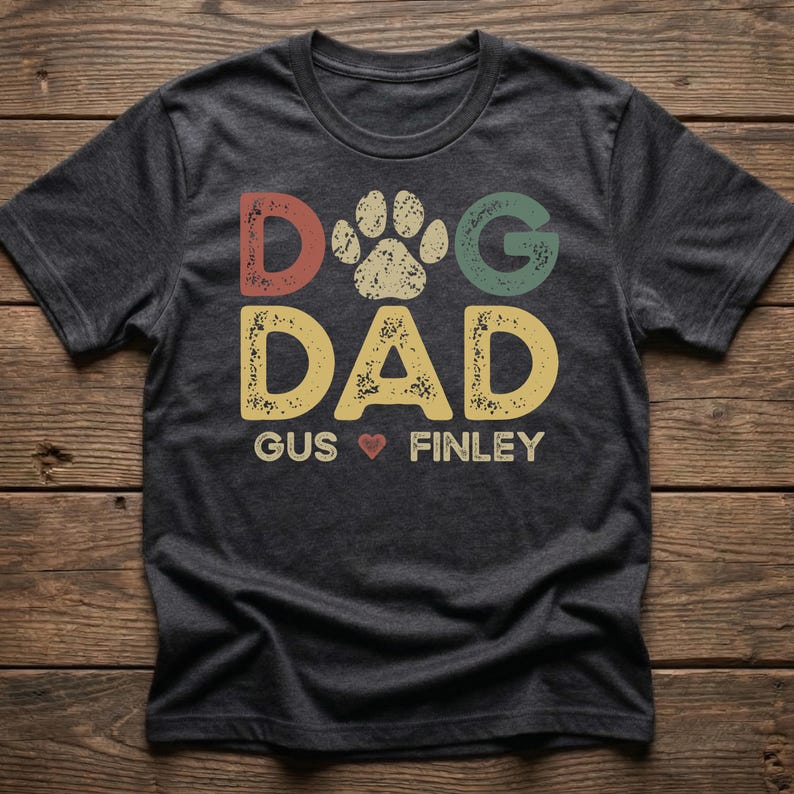 May include: Dark gray t-shirt with the words "DOG DAD" in colorful, distressed lettering, a paw print, and the names "Gus" and "Finley" with a heart symbol. The shirt is on a wooden surface.