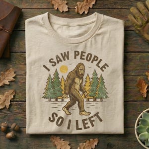 May include: Beige t-shirt with a Bigfoot graphic. The design features a walking Bigfoot in front of pine trees and a sun, with the text "I SAW PEOPLE SO I LEFT" in brown lettering. The shirt is folded on a wooden surface.