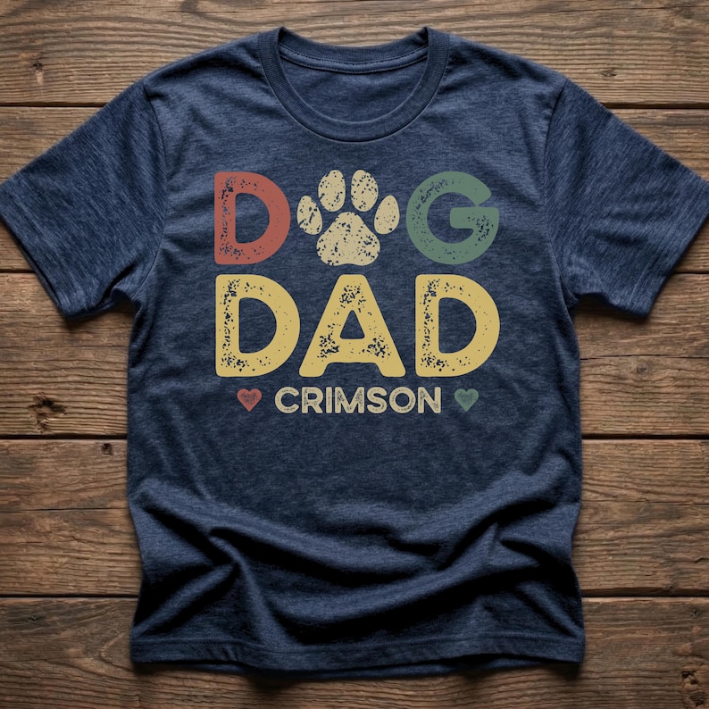 May include: Navy blue t-shirt with the words "DOG DAD" in colorful, distressed lettering. A paw print graphic is above the word "DAD". The word "CRIMSON" is below the words "DOG DAD", with a heart on each side. The shirt is on a wooden background.