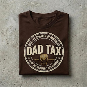 Puede incluir: Una camiseta marr&oacute;n doblada con un gr&aacute;fico circular que dice "Quality Control Department DAD TAX Certified Sampling Required. 1 Bite Minimum." El gr&aacute;fico incluye un dibujo de patatas fritas.