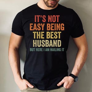 May include: Black t-shirt with the text "IT'S NOT EASY BEING THE BEST HUSBAND BUT HERE I AM NAILING IT" in red, orange, yellow, and white. The shirt has a vintage, distressed look.