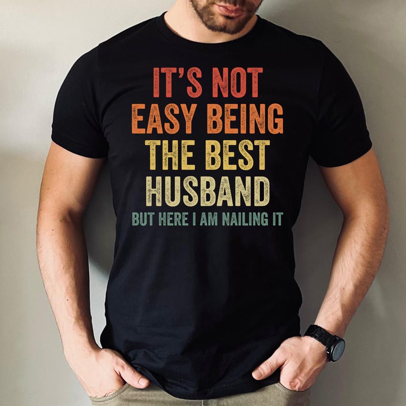 May include: Black t-shirt with the text "IT'S NOT EASY BEING THE BEST HUSBAND BUT HERE I AM NAILING IT" in red, orange, yellow, and white. The shirt has a vintage, distressed look.