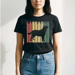 May include: Black t-shirt featuring a Labrador silhouette with the word "LABRADOR" above it. The design includes vertical stripes in red, yellow, and green. The shirt is made of a soft material.