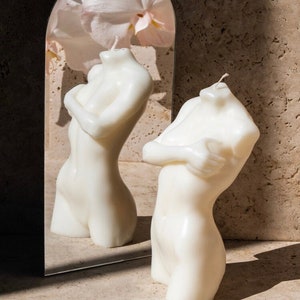 May include: Two white, sculpted candles in the shape of a human torso. The candles are placed on a light brown surface in front of a mirror. The mirror is arched and reflects a pink flower.