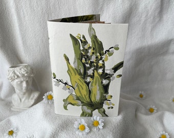 May include: A white notebook with a green and white floral design. The flowers are stylized and resemble lilies. The notebook is closed and sitting on a white surface.