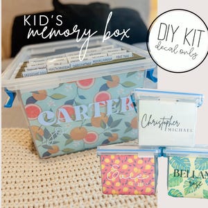 May include: A clear plastic storage box with a colorful citrus fruit design and the name "Carter" on the front. The box is labeled "Kid's memory box" and is being used to store smaller clear plastic boxes with personalized labels. The labels include the names "Christopher Michael", "Ella Claire", and "Bellamy".
