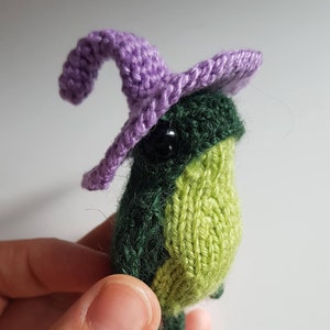 Op de afbeelding: Een kleine groene gebreide kikker met een paarse gebreide heksenhoed. De kikker heeft een groen lichaam en een lichtgroene buik.