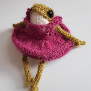 Op de afbeelding: Een gebreide kikker die een roze jurk draagt met een witte strik. De kikker heeft groene ogen en zit met gekruiste benen.