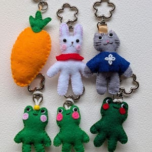 Puede incluir: Un conjunto de seis llaveros de fieltro que incluyen una zanahoria naranja brillante, un conejo blanco con bufanda roja, un gato gris con camisa azul y tres ranas verdes con mejillas rosadas, unidos a anillas metálicas plateadas en forma de flor.