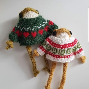 Puede incluir: Dos figuras de rana hechas a mano con pequeños suéteres de punto. Una rana tiene un suéter verde con corazones rojos, la otra un suéter blanco con patrones rojos y verdes. Las ranas tienen patas largas y delgadas y brazos pequeños.