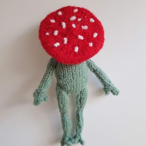 May include: A knitted frog toy wearing a red mushroom hat with white spots. The frog is green and has a long body with four legs.