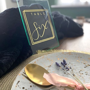 Puede incluir: Un letrero de número de mesa negro y dorado con el texto "TABLE Six" sobre un fondo de vidrio transparente. El letrero está sobre una superficie de tela negra.