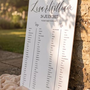 May include: A white rectangular wedding seating chart sign with black text reading Lisa & William, 24 Juin 2023, Please Take Your Seat, and lists of names under columns labeled One and Two, accented by a single cream-colored rose at the base.