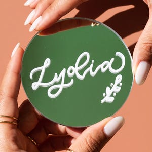 May include: A round, dark green compact mirror featuring the name Lydia written in white cursive script across the center, accompanied by a small white floral graphic element positioned in the lower right corner of the circular surface.