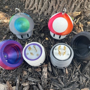 May include: Five different 3D printed mushroom-shaped objects.  Two are red and white, one is black, one is purple and pink, and one is gray and green.  Each mushroom has a different design on the top, including a standard electrical outlet, a blank surface, and a circular design.