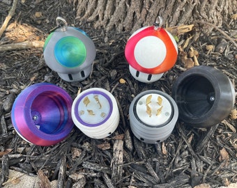 May include: Five different 3D printed mushroom-shaped objects.  Two are red and white, one is black, one is purple and pink, and one is gray and green.  Each mushroom has a different design on the top, including a standard electrical outlet, a blank surface, and a circular design.