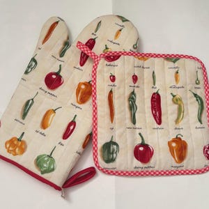 Könnte beinhalten: Ein gestepptes Ofenhandschuh- und Topflappen-Set. Der cremefarbene Stoff zeigt Illustrationen verschiedener Chilischoten mit ihren Namen. Die Kanten sind mit rotem Stoff eingefasst. Der Topflappen hat einen rot-weiß karierten Rand.