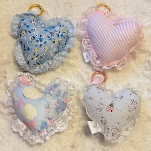May include: Four heart-shaped fabric ornaments with lace trim. One is blue with floral print, one is pink and white striped, one is light blue with a fruit pattern, and one is light blue with a small floral print. Each has a gold-tone metal ring.
