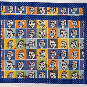 May include: Blue fabric placemat with a repeating pattern of a cartoon character's face. The character has a blue, yellow, and red face with a wide smile and large eyes. The placemat has a blue border with white polka dots.