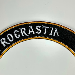 May include: Black, arched patch with the word "PROCRATIN" embroidered in white. The patch has a gold border and white stitching. A humorous accessory for clothing or bags.