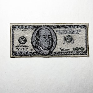 May include: Embroidered patch depicting a United States $100 bill. The patch features a portrait of Benjamin Franklin, the text "The United States of America", and the number "100". The patch is rectangular with a detailed design.