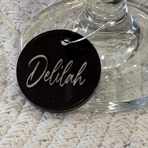 May include: A black, round wine glass charm with the name "Delilah" in white script. The charm is attached to a clear wine glass. The background is a textured, off-white surface.