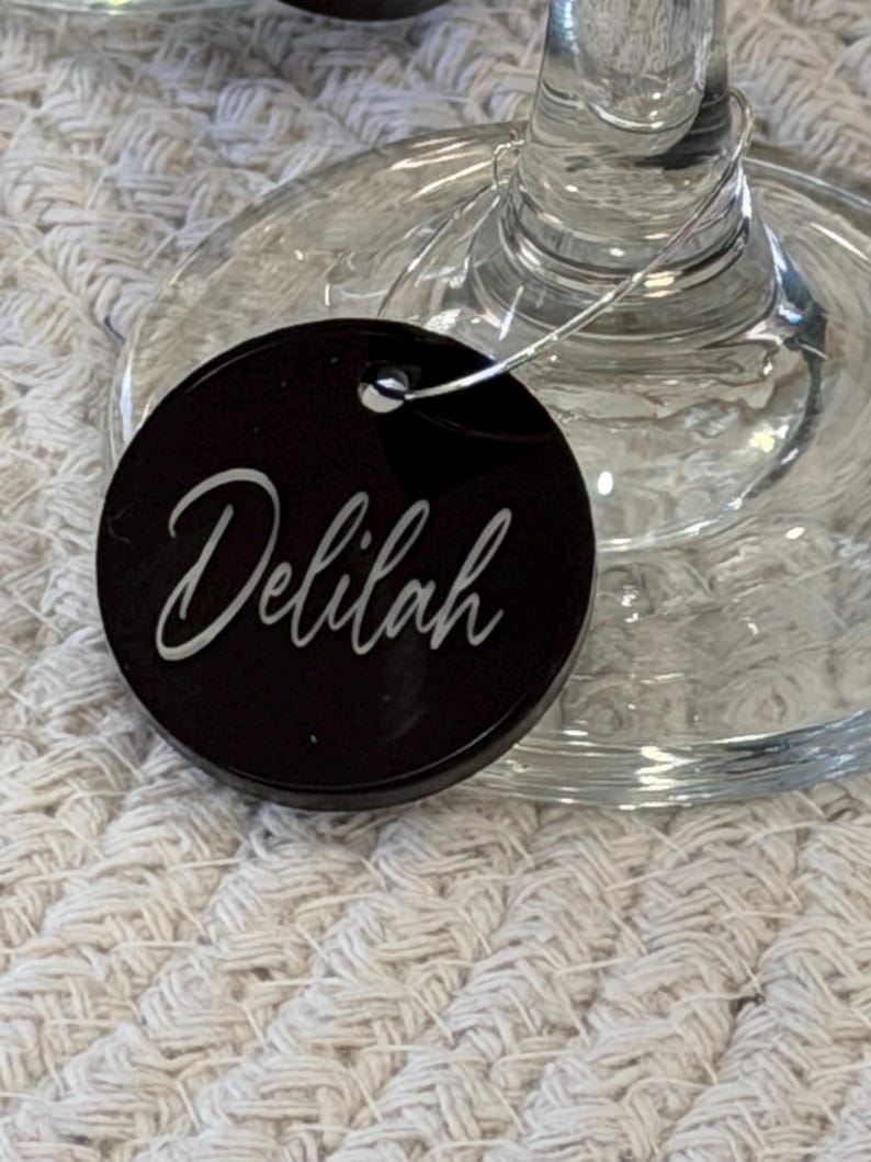 May include: A black, round wine glass charm with the name "Delilah" in white script. The charm is attached to a clear wine glass. The background is a textured, off-white surface.